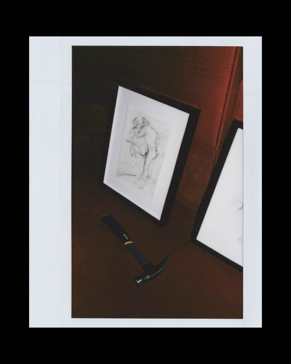 A framed sketch of a seated nude figure resting against a dark wall with a hammer placed nearby.