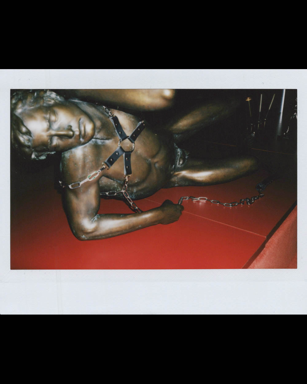 A bronze statue of a reclining male figure wearing a harness and chained wrist on a red surface.
