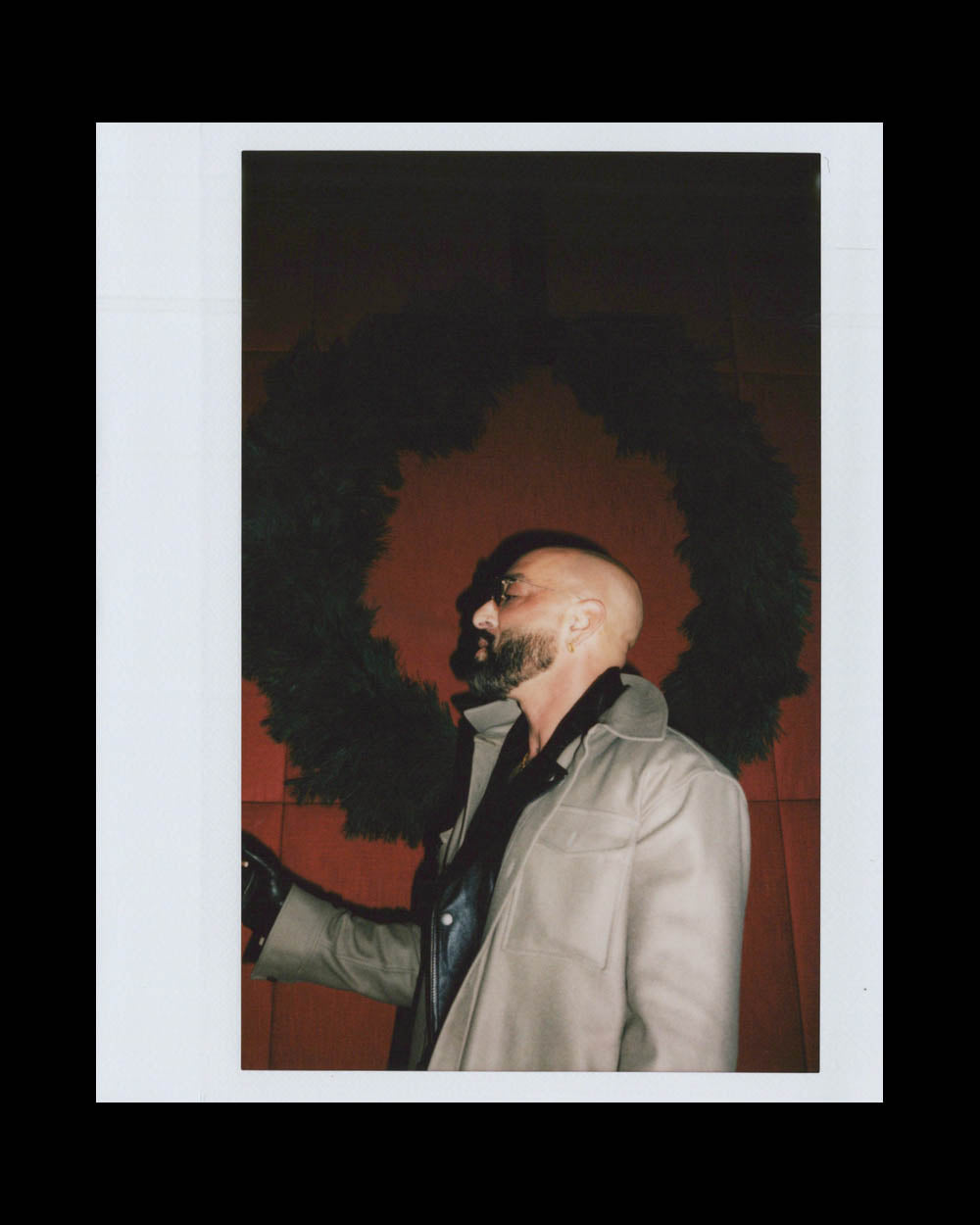 A bald man with a beard wearing glasses and a beige coat stands in front of a large dark wreath on a red wall.
