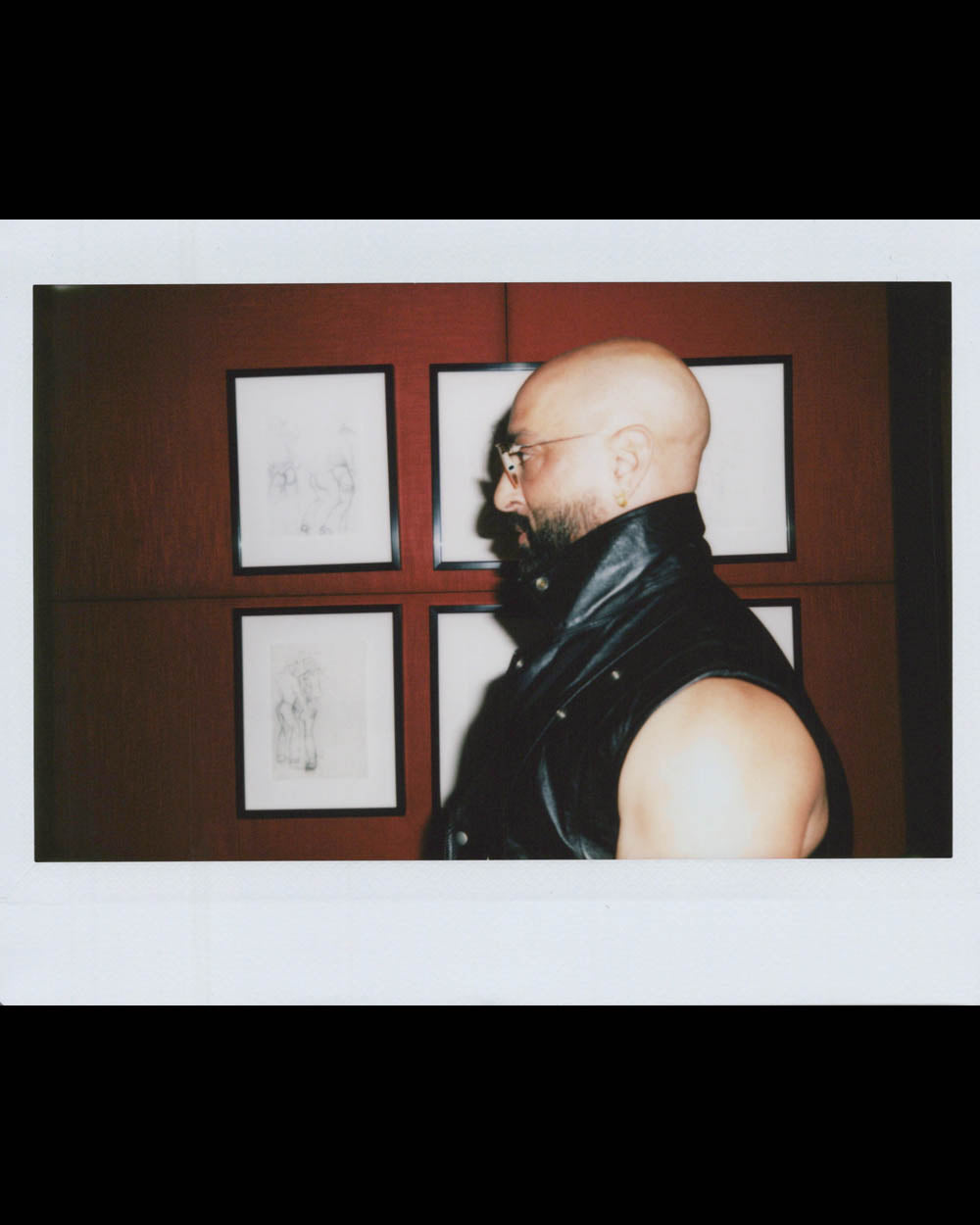 A bald man with glasses and a beard wearing a sleeveless leather vest stands in profile by framed sketches on a red wall.