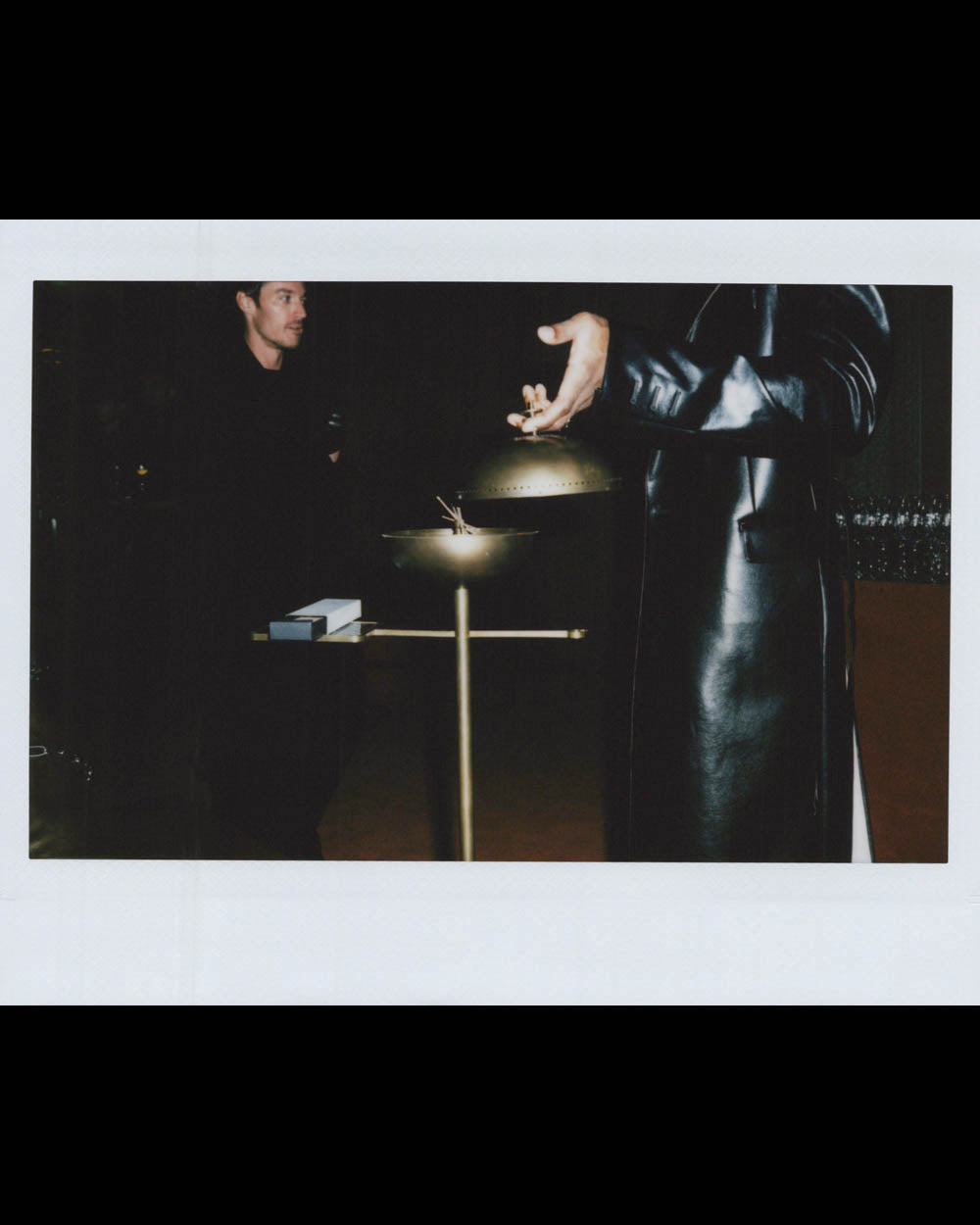 A person in a black leather coat gestures near a metallic stand while another man watches in dim lighting.