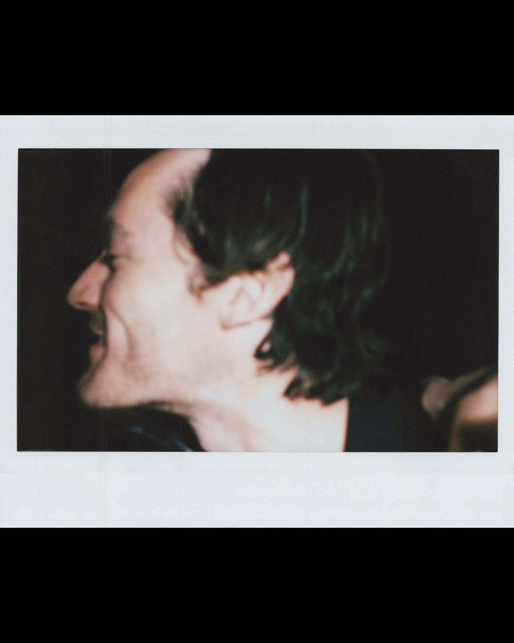 A close-up side profile of a smiling man with dark hair against a black background.