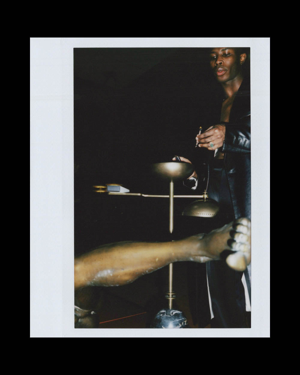 A man in a black leather jacket holds a lit cigarette near a bronze sculpture with an outstretched leg.