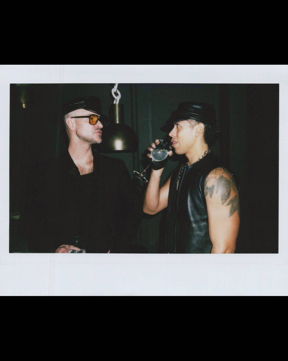 Two men in black leather outfits and hats engage in conversation in a dimly lit indoor setting.
