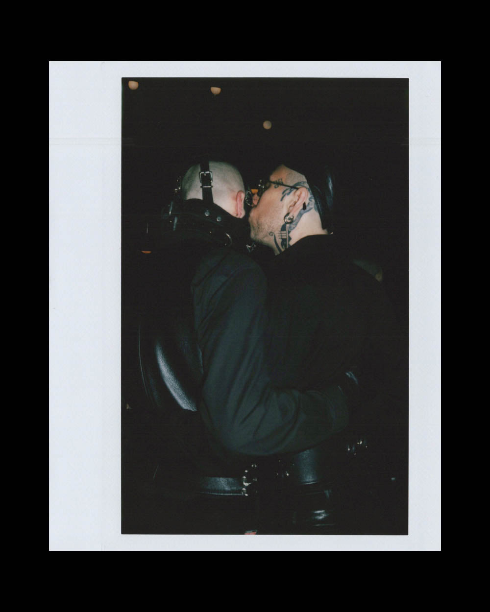 Two men dressed in black leather, one with visible tattoos, share a close moment in a dark setting.