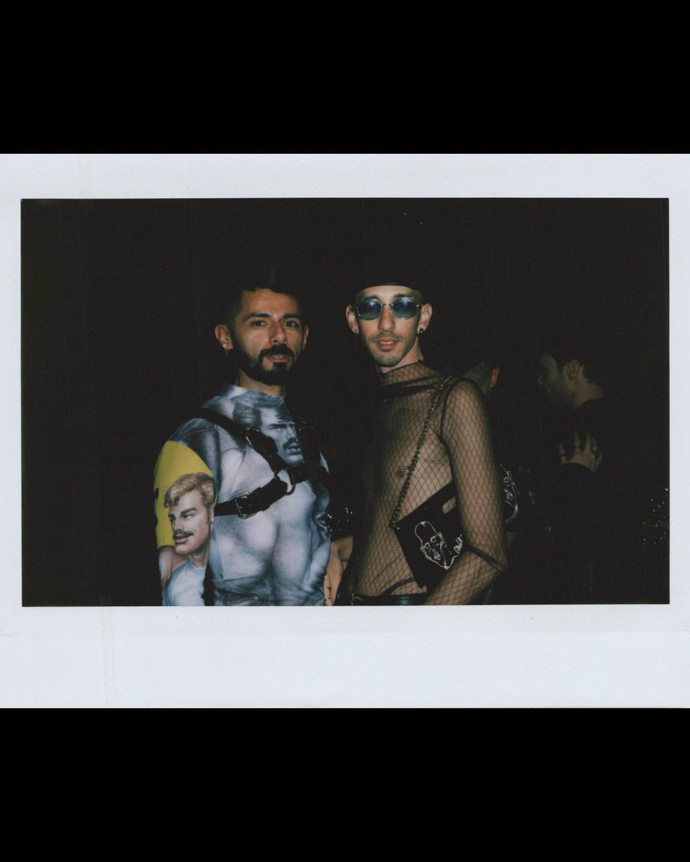 Two men pose in low light, one wearing a detailed graphic top, the other in a black mesh shirt and tinted glasses.