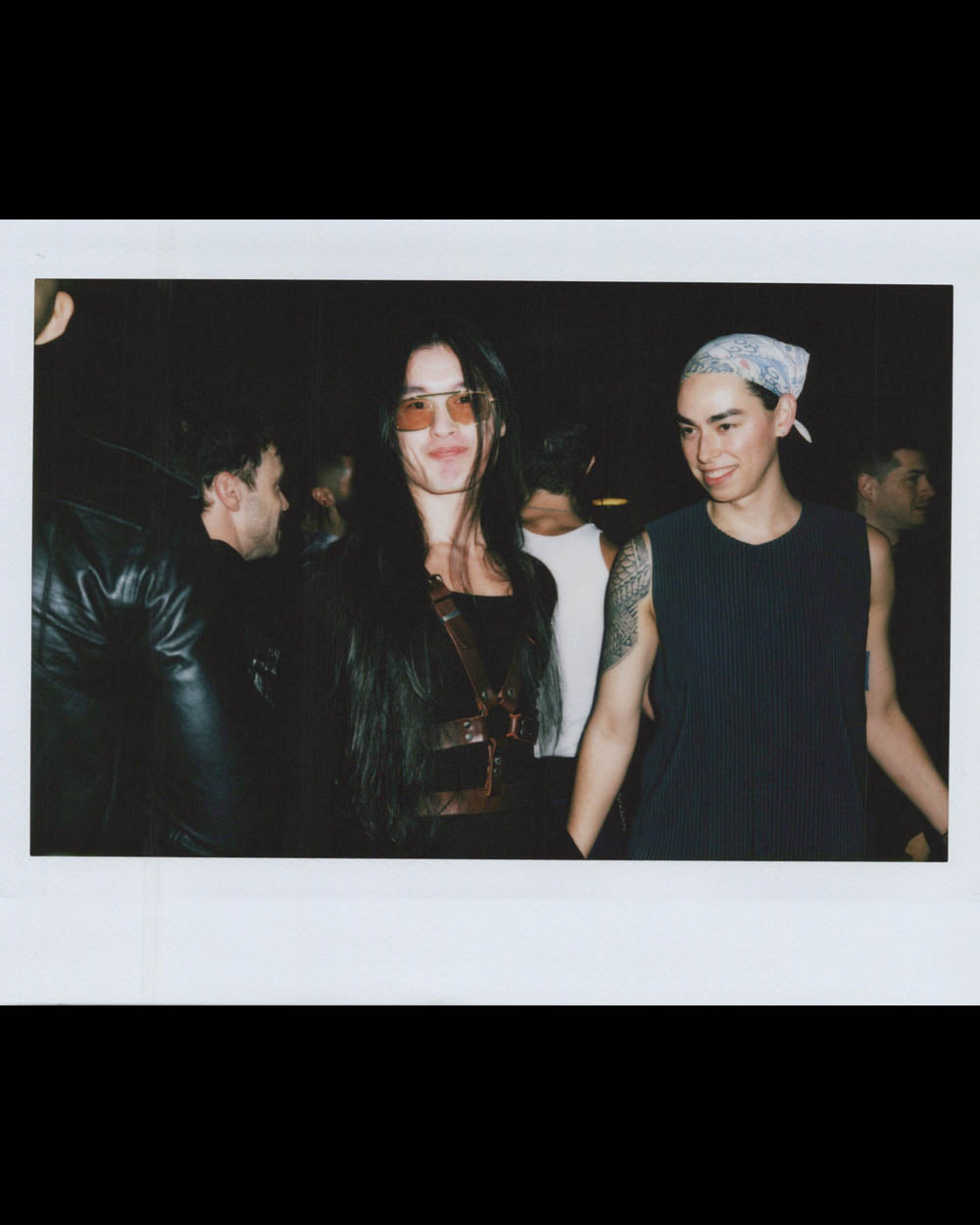 A woman with long dark hair and tinted glasses stands beside a man with a bandana and sleeveless top, both smiling.