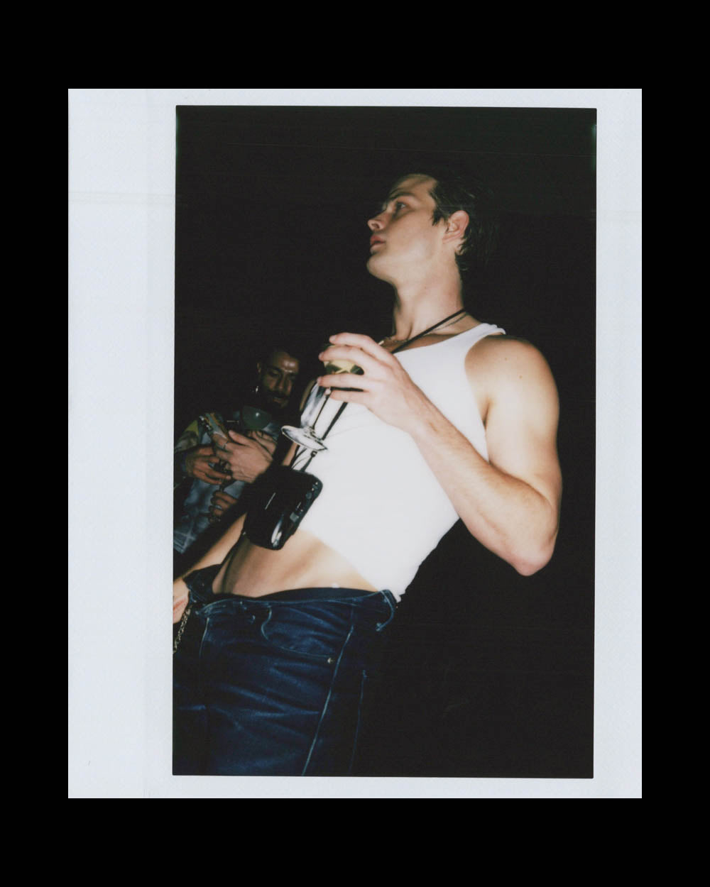 A young man in a white tank top and dark pants holds a glass, standing against a dark background.