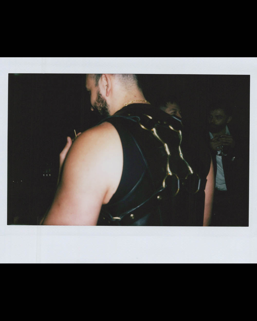 A man wearing a sleeveless black outfit with metallic chain details on the back in a dark setting.