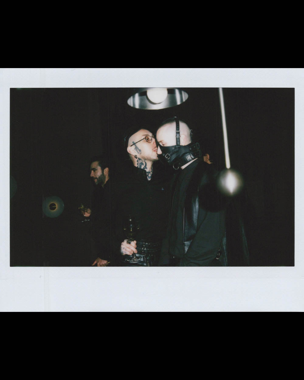 Two men dressed in black, one wearing tattoos and glasses, the other with a leather mask, embrace in a dim room.
