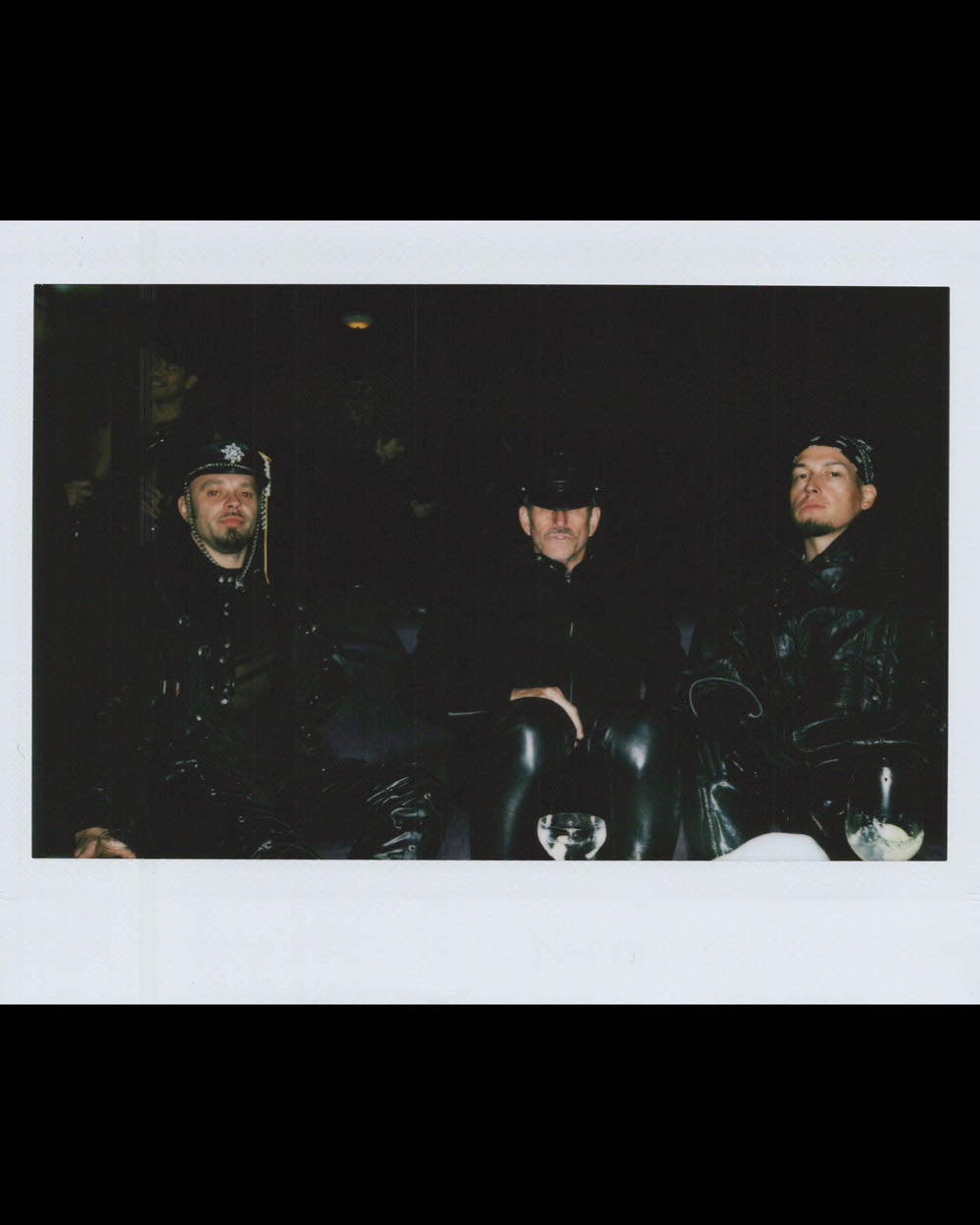 Three men dressed in dark leather and hats sitting closely together in a low-lit setting with glasses in front.