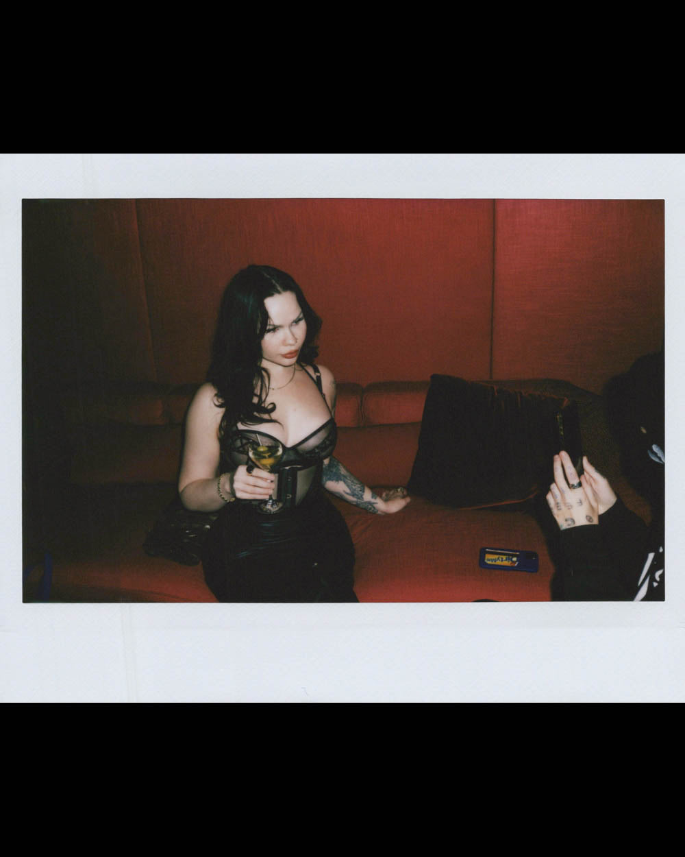 A woman with long dark hair and tattoos sits on a red couch holding a cocktail glass in a dim room.