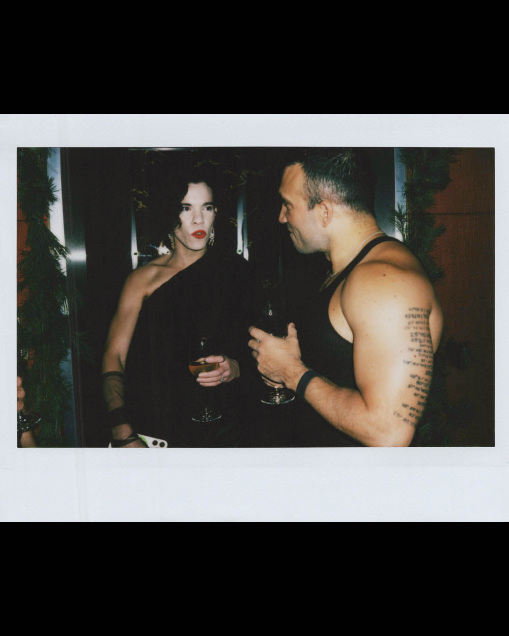 A person with dark curly hair wearing a one-shoulder black outfit talks to a muscular man in a black tank top.