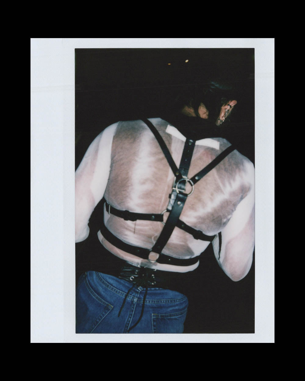 A shirtless person wearing a black leather harness and blue jeans seen from the back in a dark setting.
