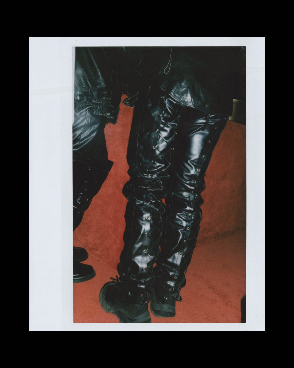 Close-up of two people wearing black leather pants and boots standing on a reddish floor against a dark background.