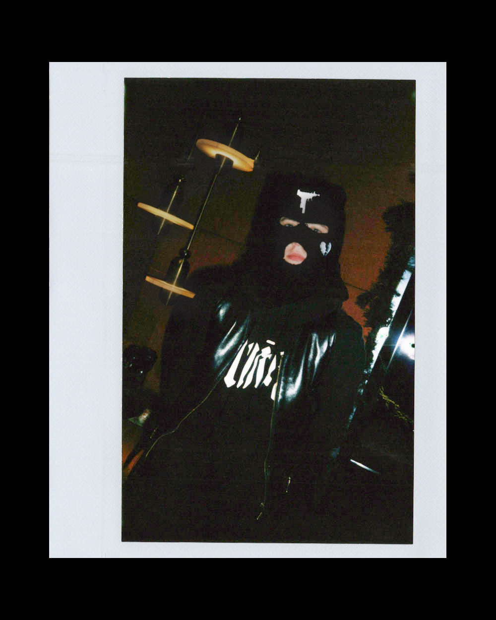 Person wearing a black leather jacket and balaclava with graphic design in a dimly lit indoor setting