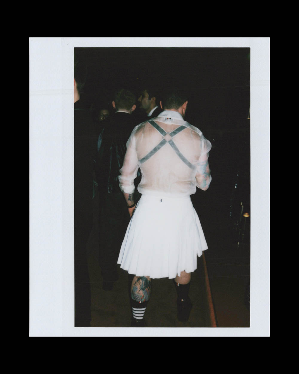 Person wearing a sheer white shirt with black harness and a white pleated skirt walking away in a dark setting.