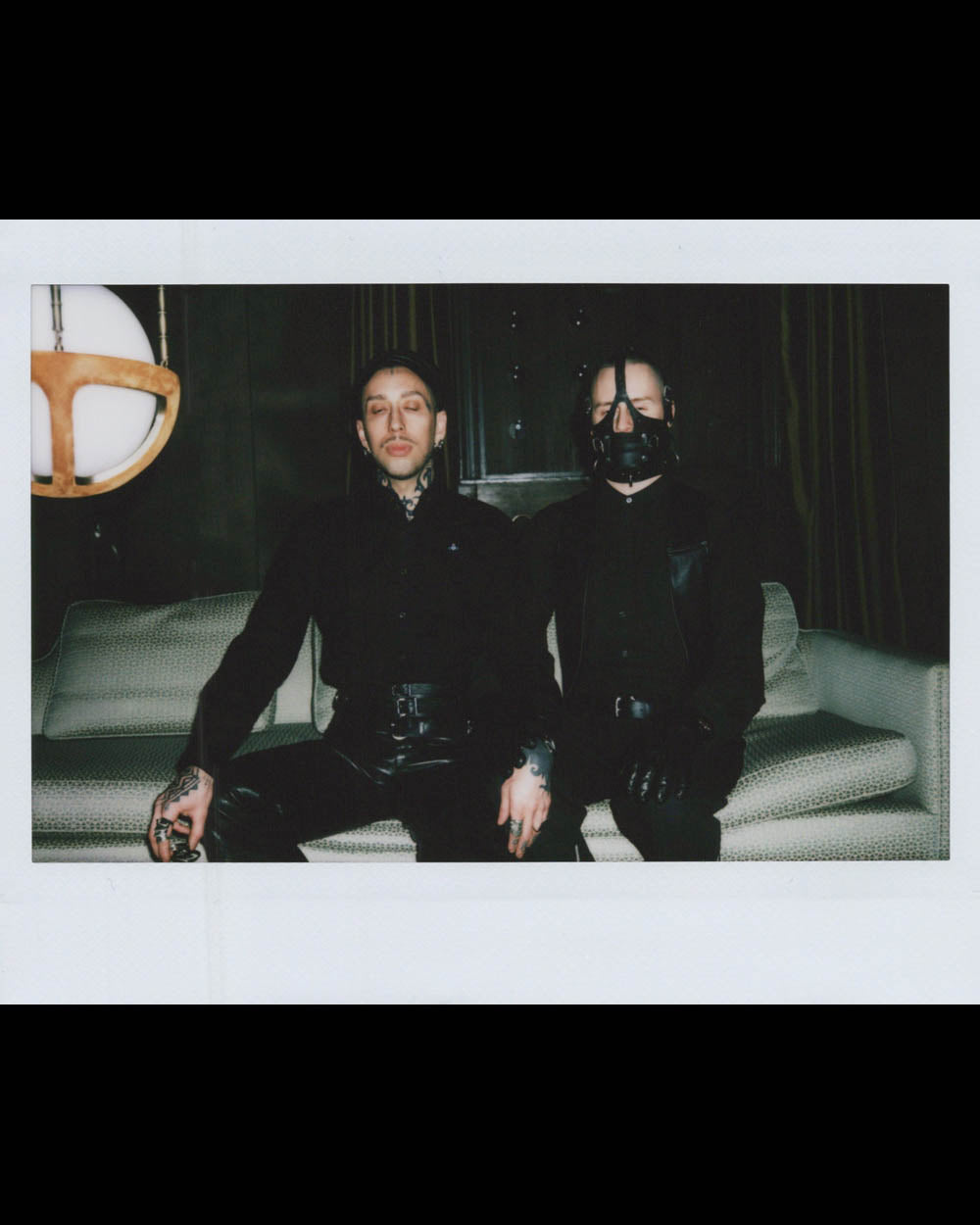 Two people dressed in black sit on a light-colored sofa in a dimly lit room, one wearing a mask covering the lower face.