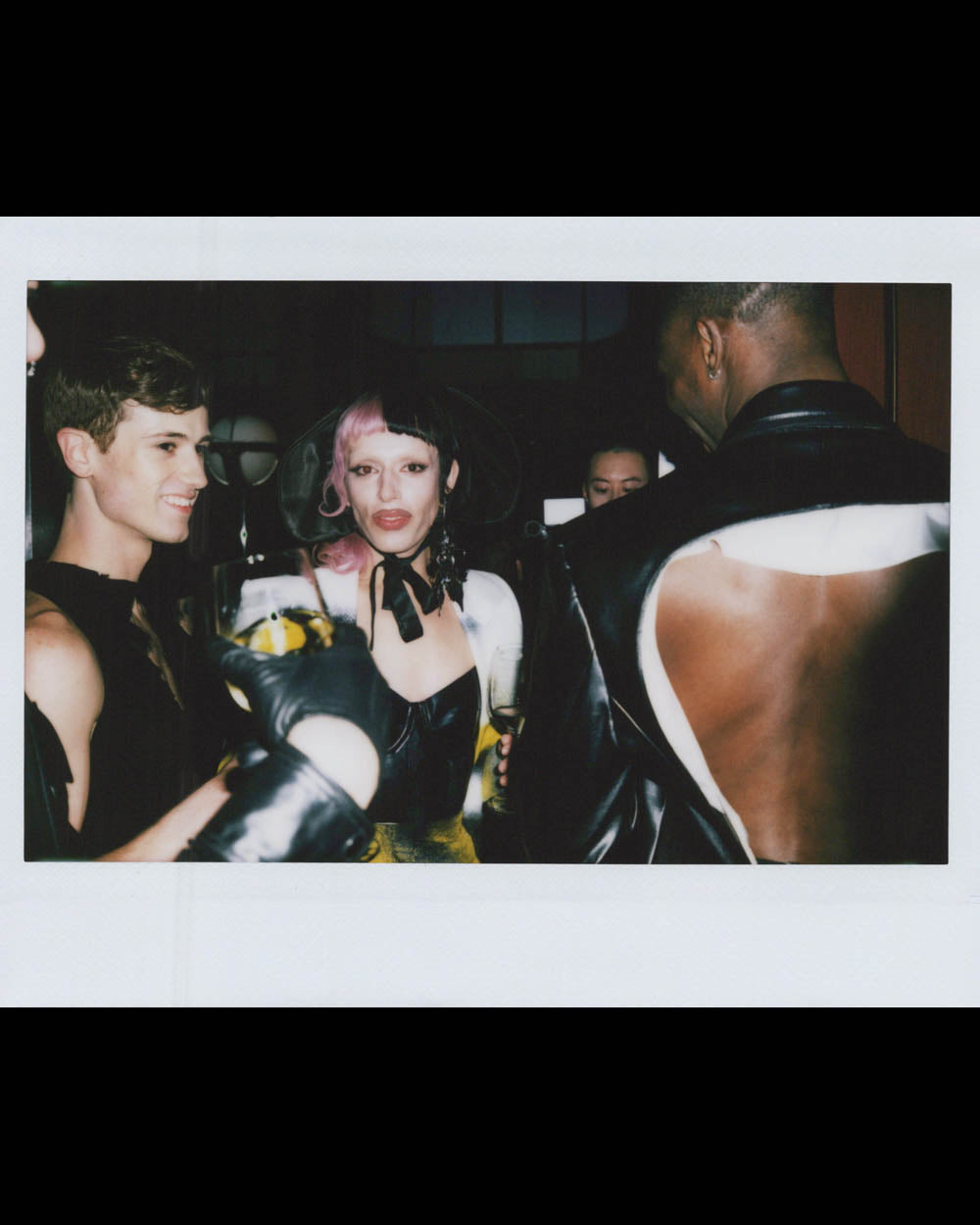 A group of people in stylish dark and white outfits socialize in a dimly lit room, one person with pink and black hair.