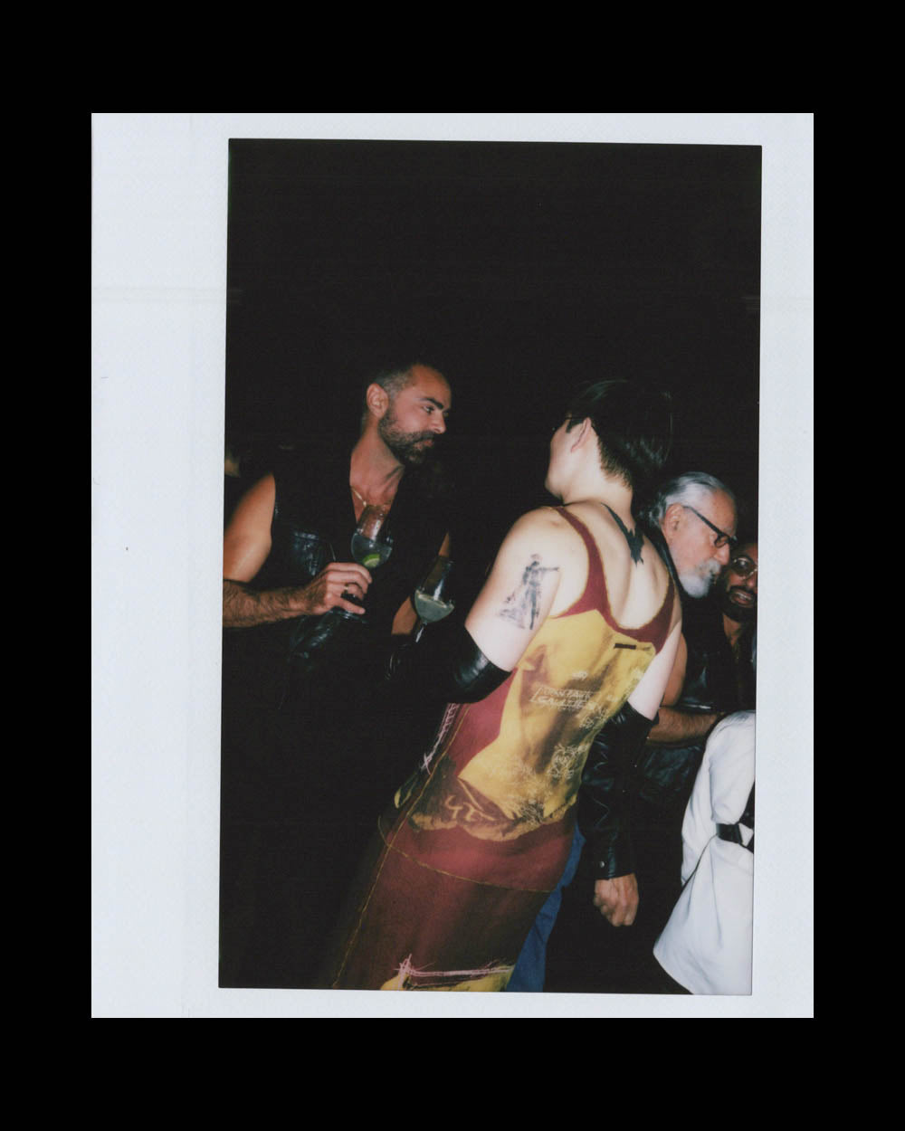 A group of people in dark clothing engage in conversation in a low-lit environment, one with a sleeveless printed dress.