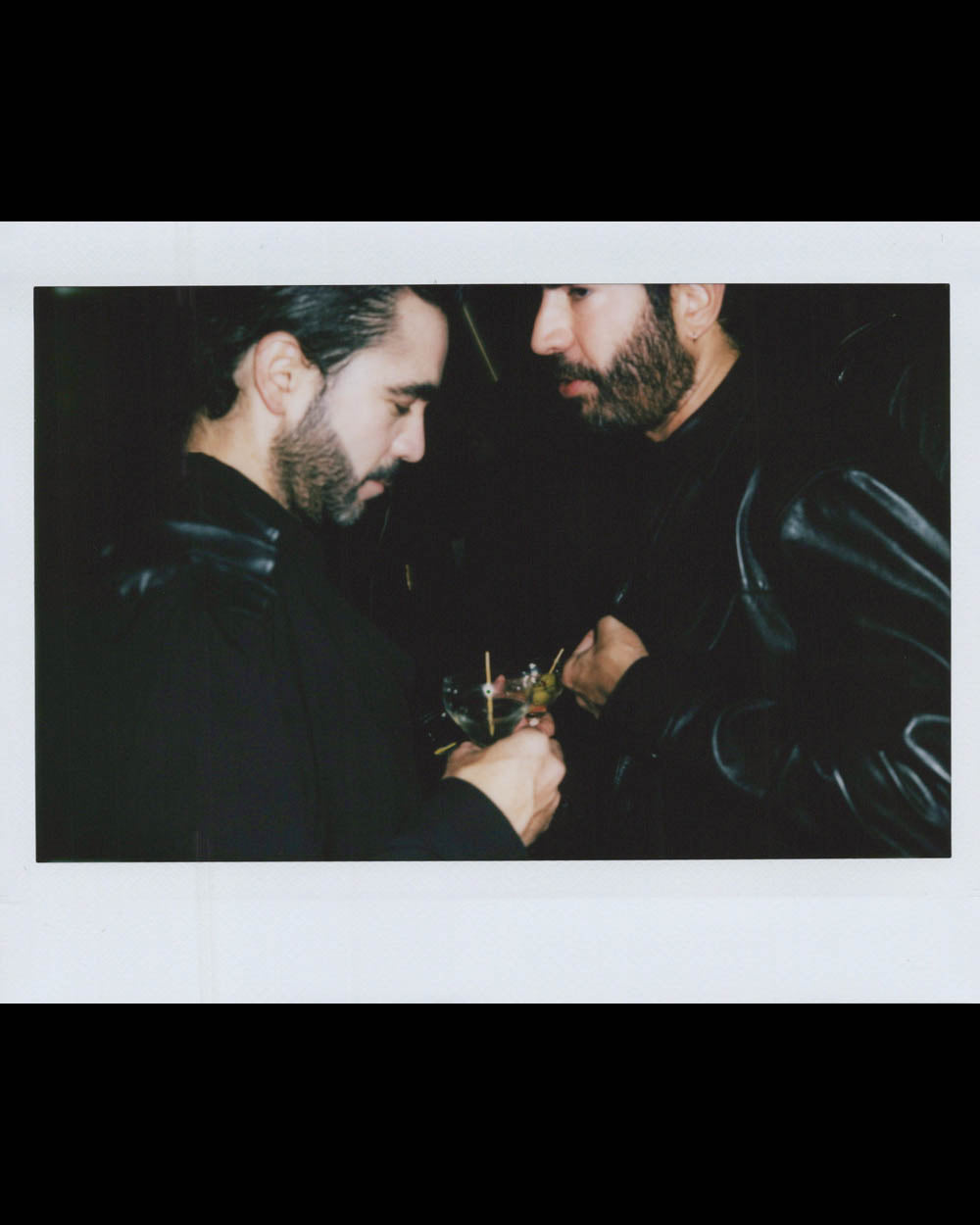Two bearded men in dark jackets share drinks and engage in conversation in a dimly lit setting.
