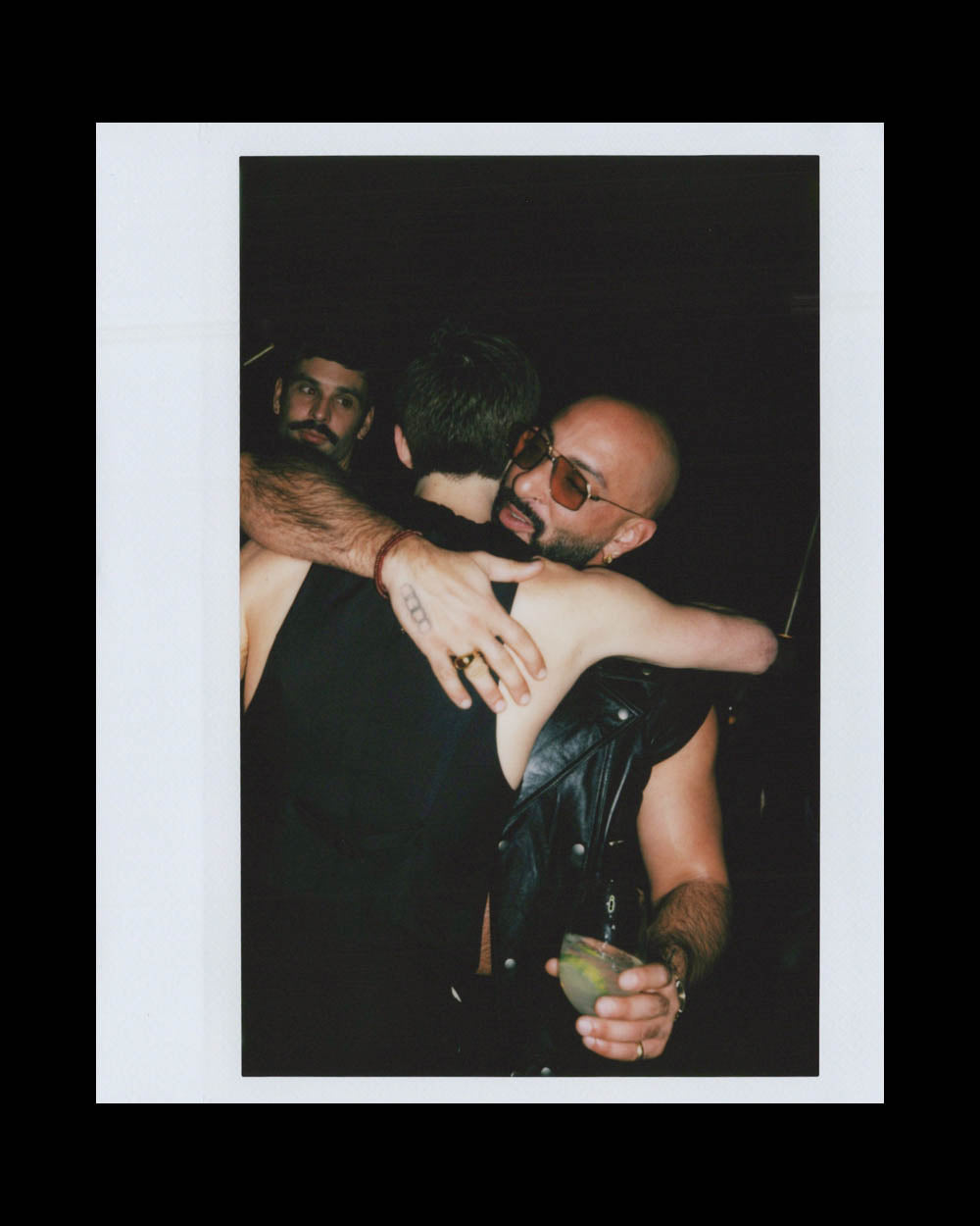 Two men embrace warmly in a dark setting, one wearing sunglasses and holding a green drink, another man nearby watches.