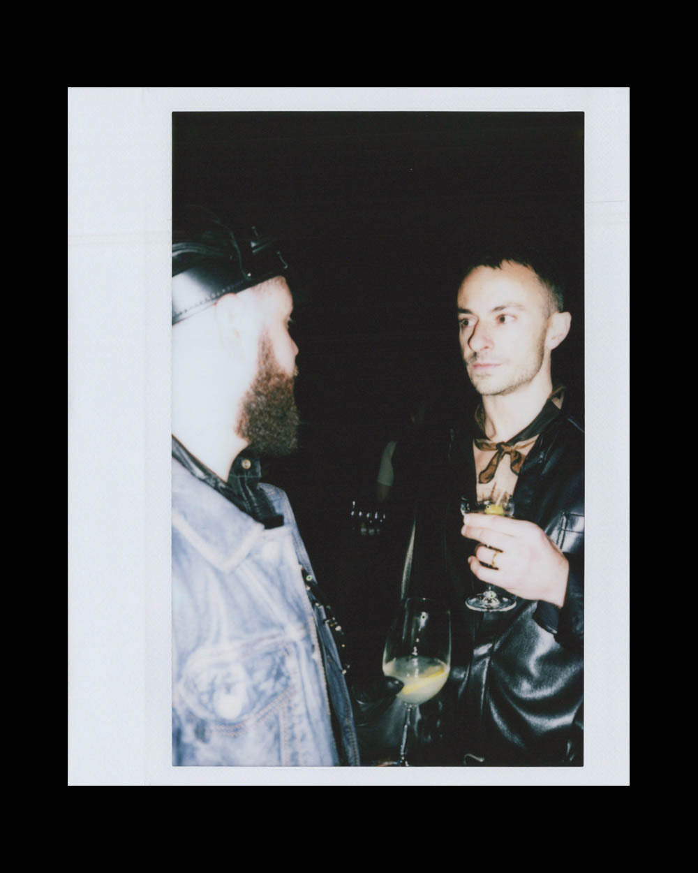 Two men in leather jackets hold drinks and converse in a dimly lit environment with a dark background.