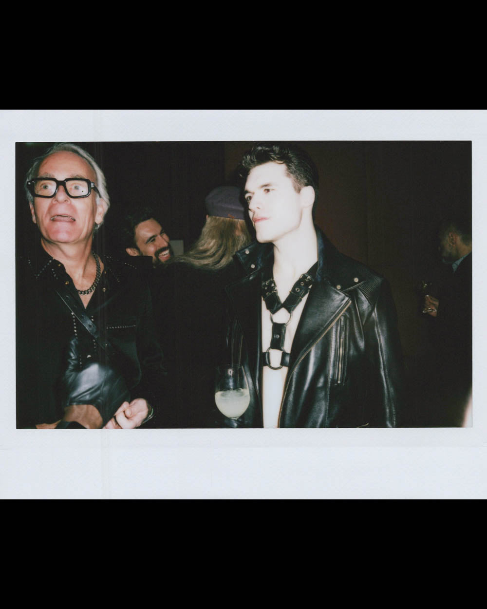 Two men in dark outfits, one wearing glasses and black gloves, and the other in a leather jacket with a harness.