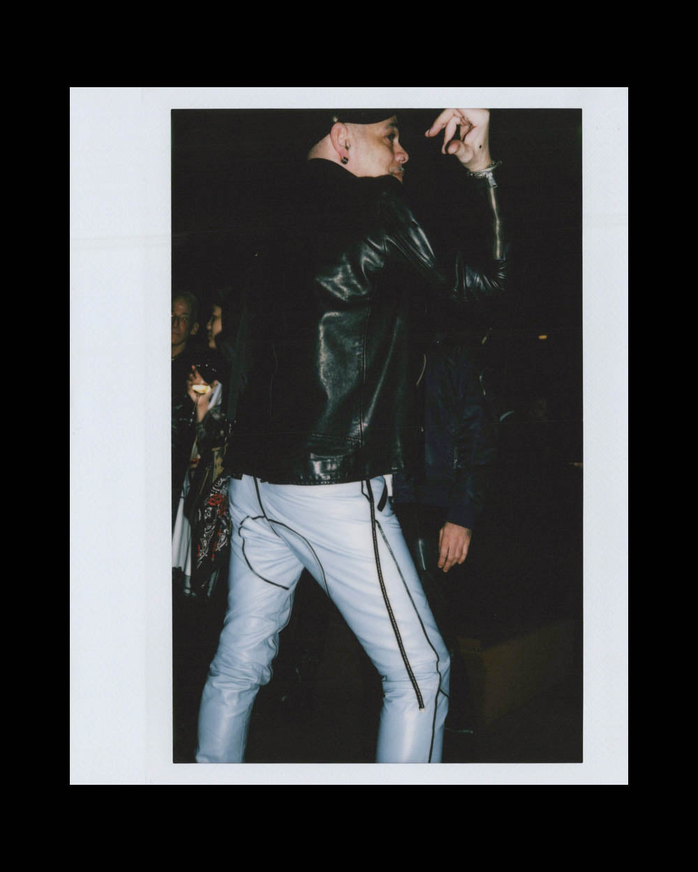 Person wearing a black leather jacket and white pants with zippers posing with arm raised in a dark setting
