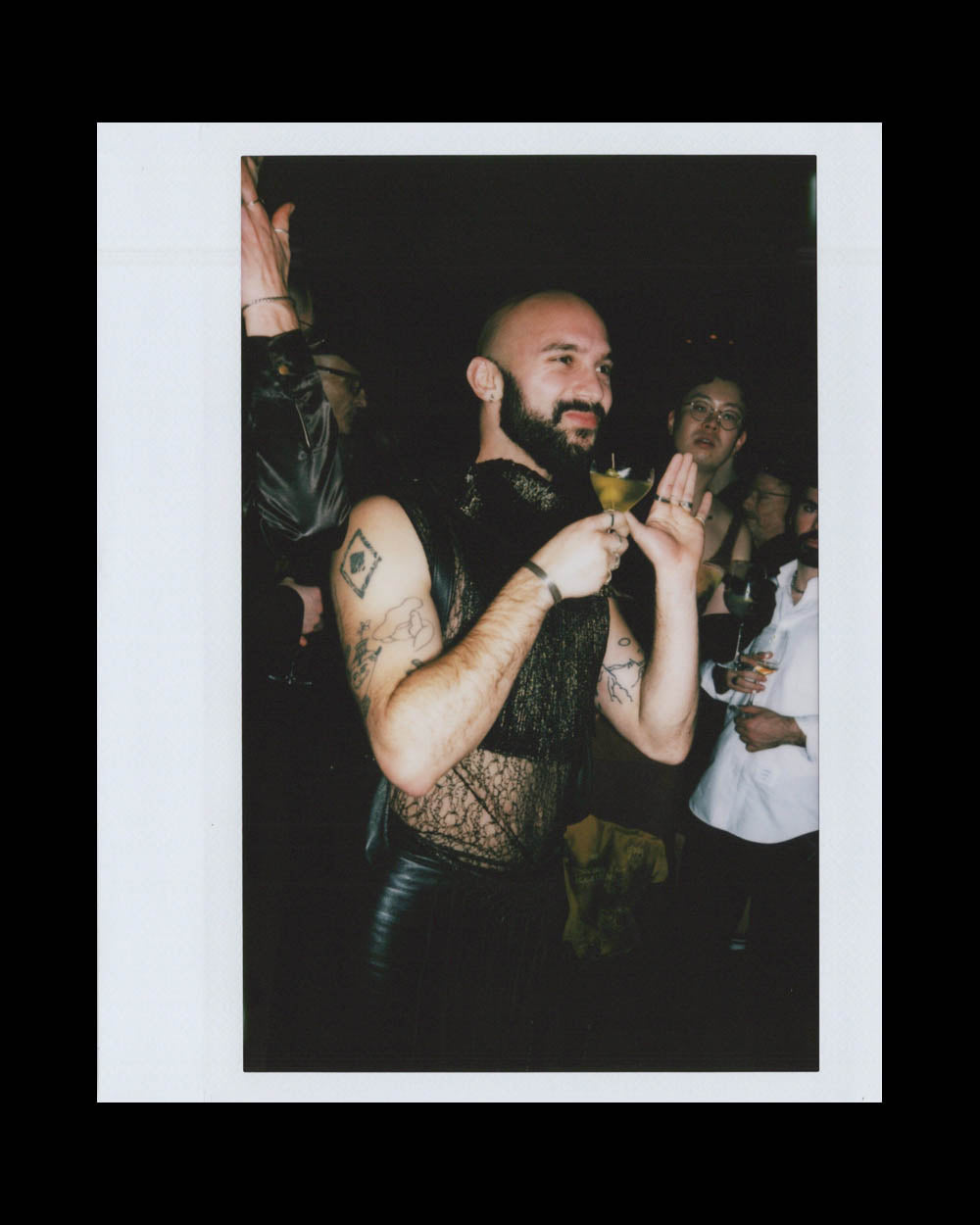 Bearded man in sleeveless mesh top and leather pants holding a cocktail and smiling at a dimly lit party.