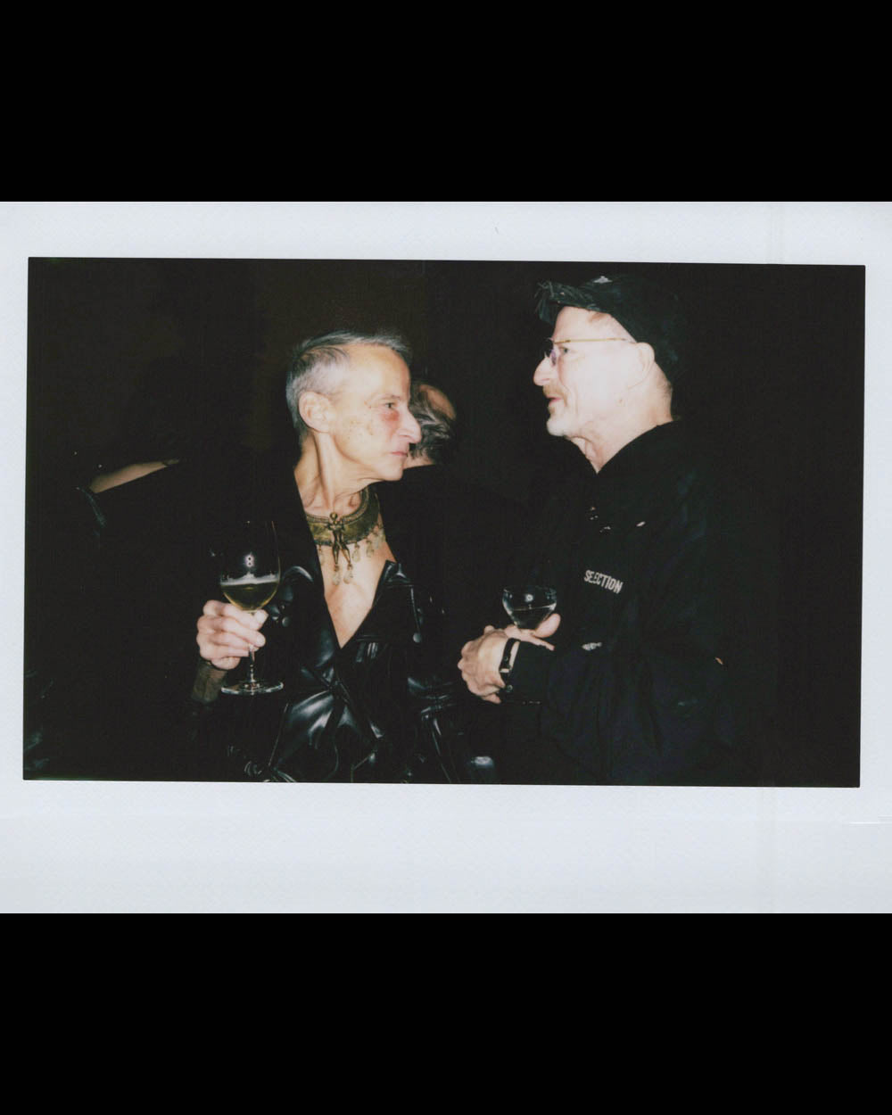 Two older men in dark clothing hold wine glasses and converse closely against a dark background.
