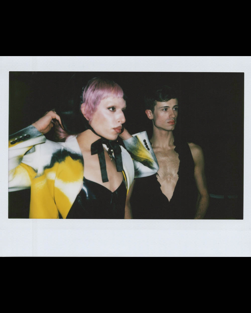 Two young people in avant-garde clothing pose against a dark background, one adjusting a neck accessory.