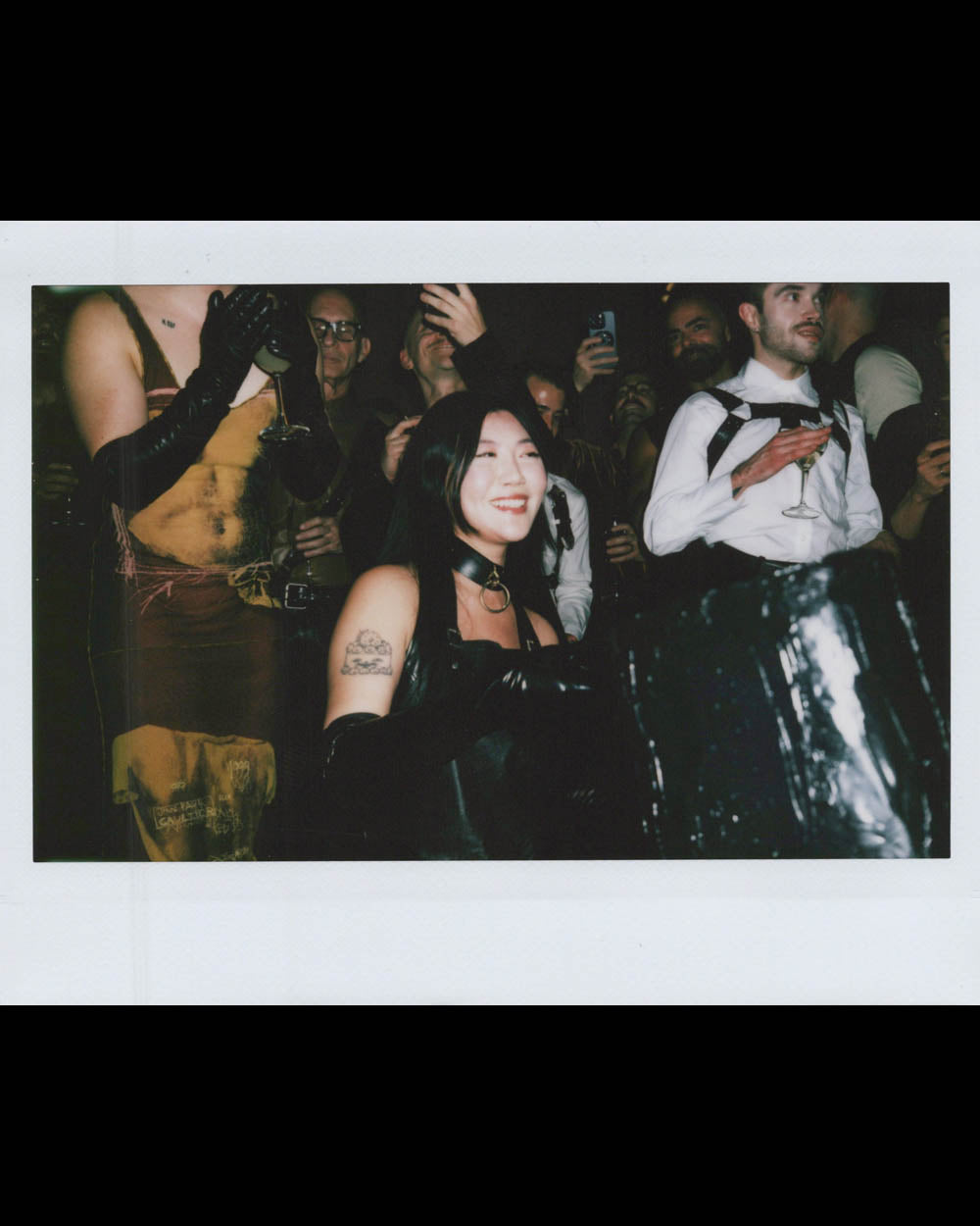 Woman wearing black gloves and choker smiles among a crowd of people dressed in dark, avant-garde attire at an event.
