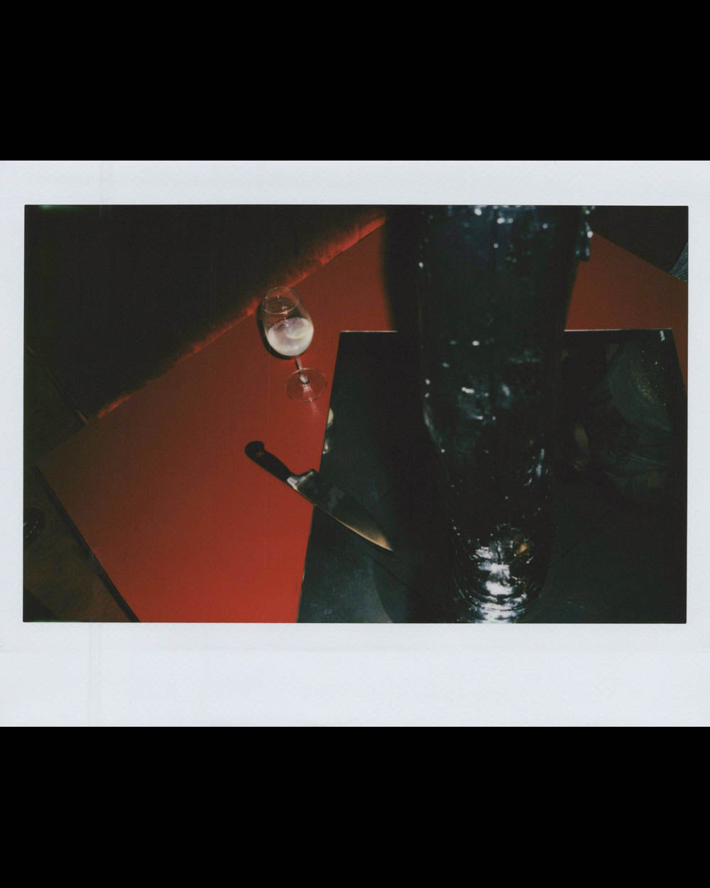 Glass of clear liquid and a knife on a reflective red and black surface with a dark, textured bottle nearby.