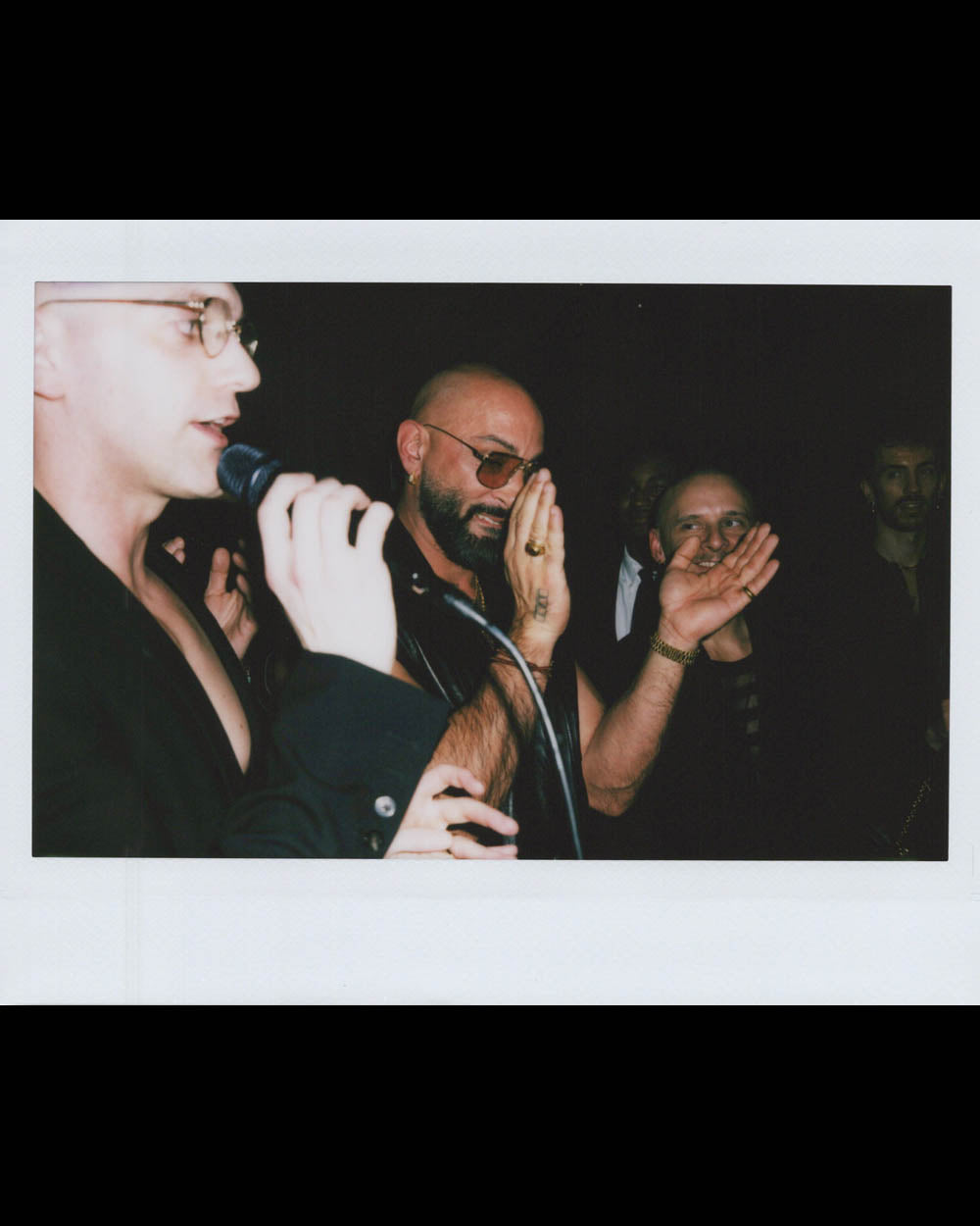 Man speaking into microphone while others behind him clap and watch in a dimly lit setting