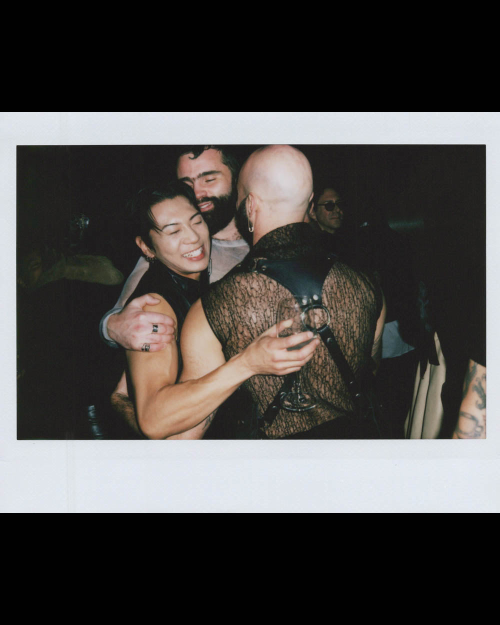 Three people embrace warmly, one holding a glass, in a dimly lit setting with others blurred in the background.