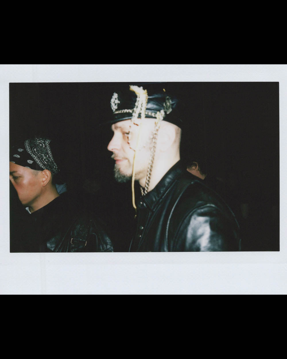 Man in leather jacket and decorative leather cap with chains stands beside person wearing bandana in dark setting