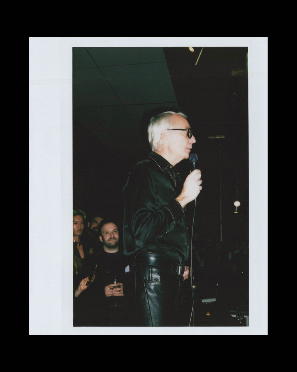 Older man in black leather shirt and pants speaking into microphone in a dark room with people watching behind him