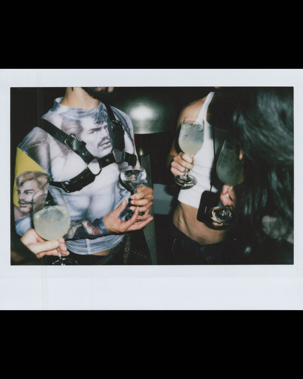 People holding glasses with light-colored drinks at a dimly lit social gathering, one wearing a graphic mesh shirt.
