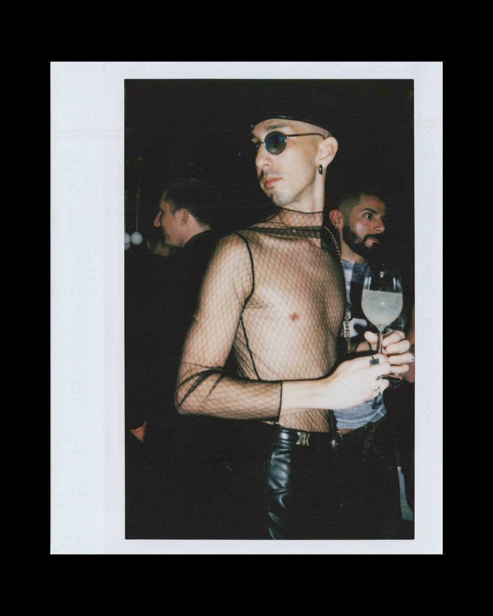 Person wearing sheer black mesh top, sunglasses, and hat, holding a drink while standing among others in low light.