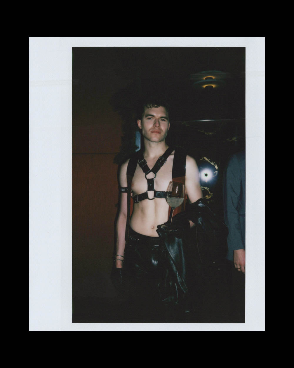 Young man wearing leather harness and pants holds a glass and jacket in dimly lit indoor setting.