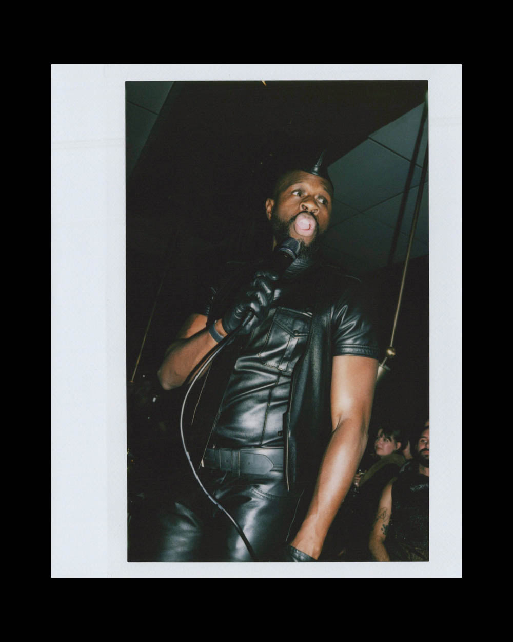 Man dressed in black leather outfit and gloves holds microphone with tongue out in a dimly lit room.