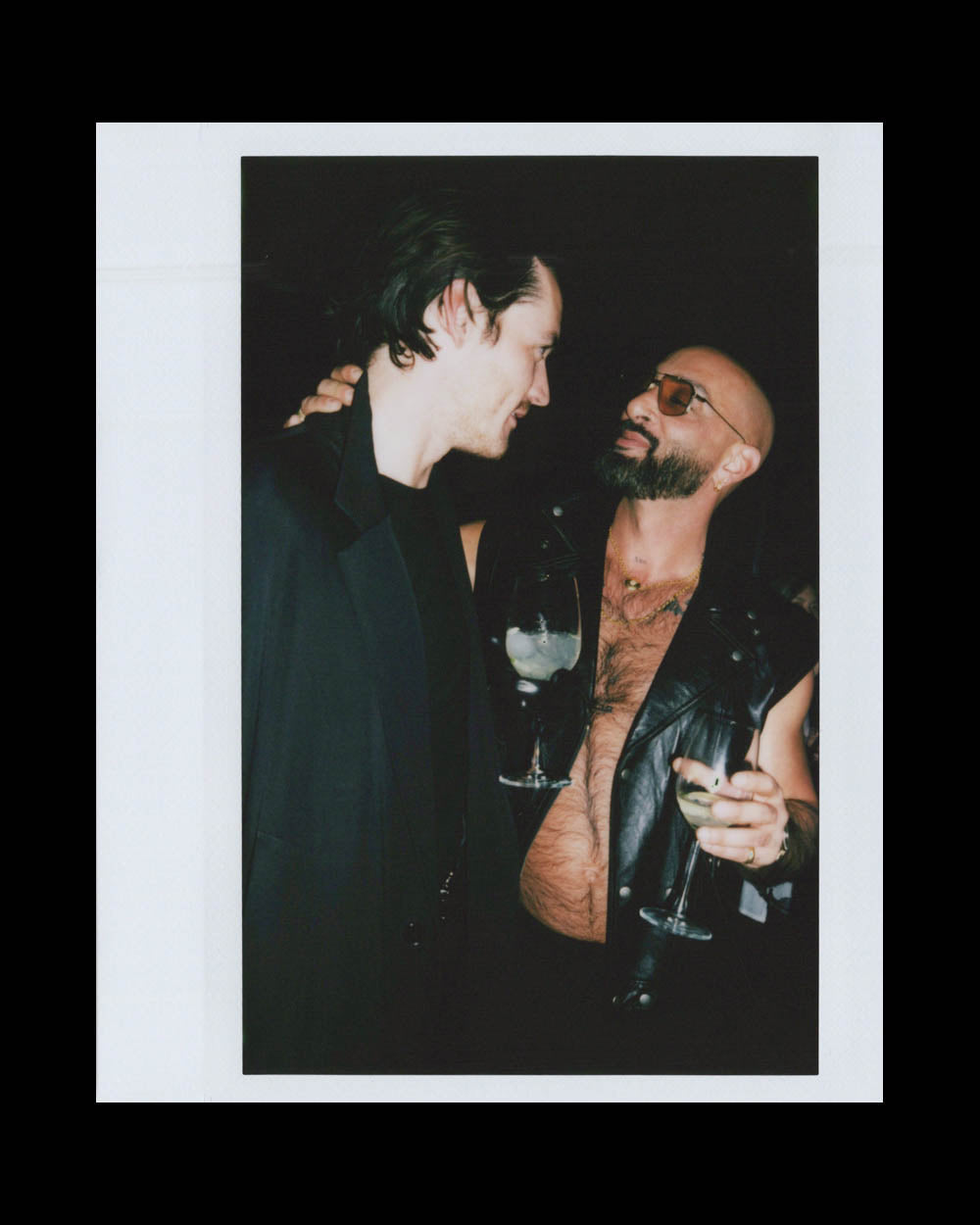 Two men facing each other sharing a moment, one wearing a sleeveless leather vest and sunglasses, holding drinks.