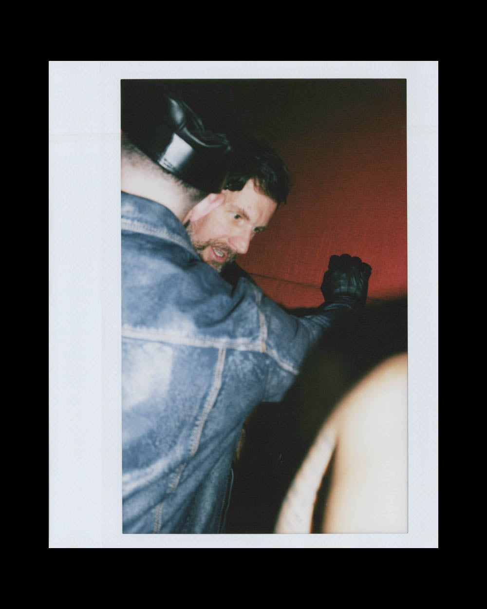 Man in denim jacket and leather cap leans close to another person wearing black leather gloves in dimly lit space.