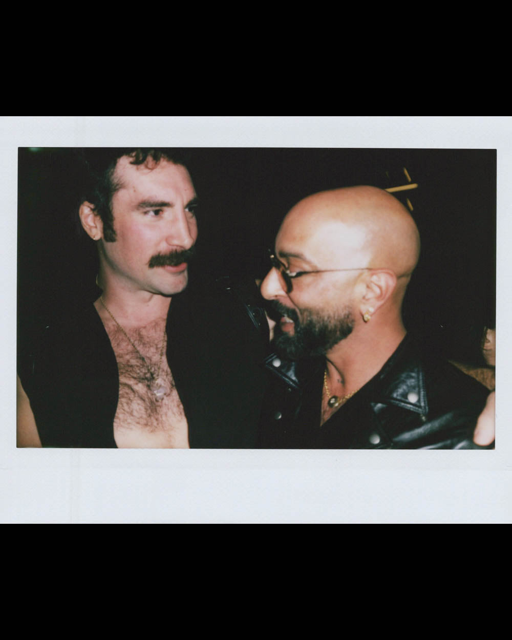 Two men close together in conversation, one with mustache and chest hair visible, the other bald with glasses and leather jacket.