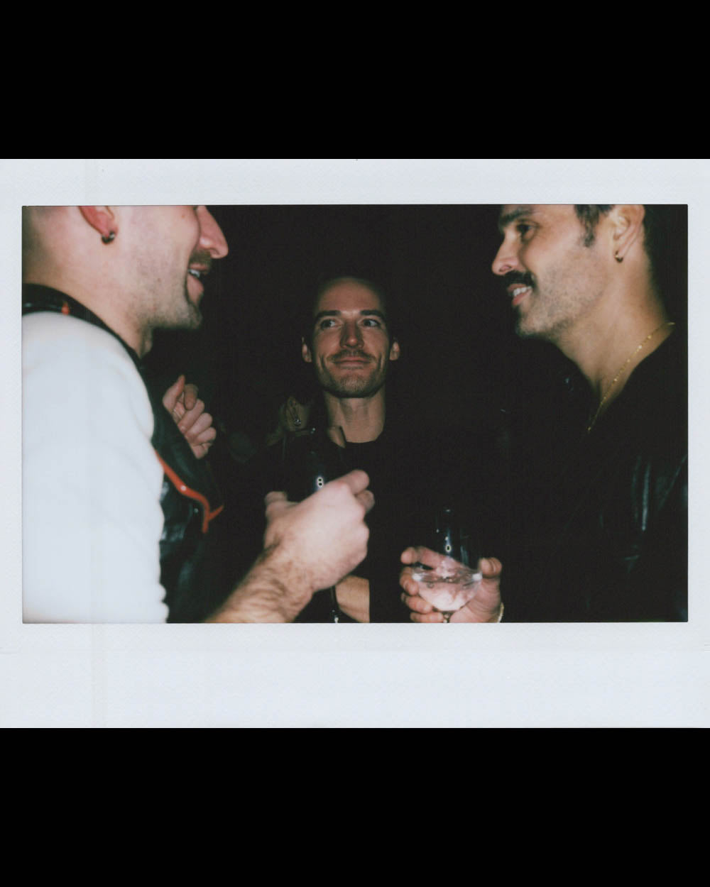 Three men in conversation at night, two holding drinks, with one smiling directly at the camera in low light.