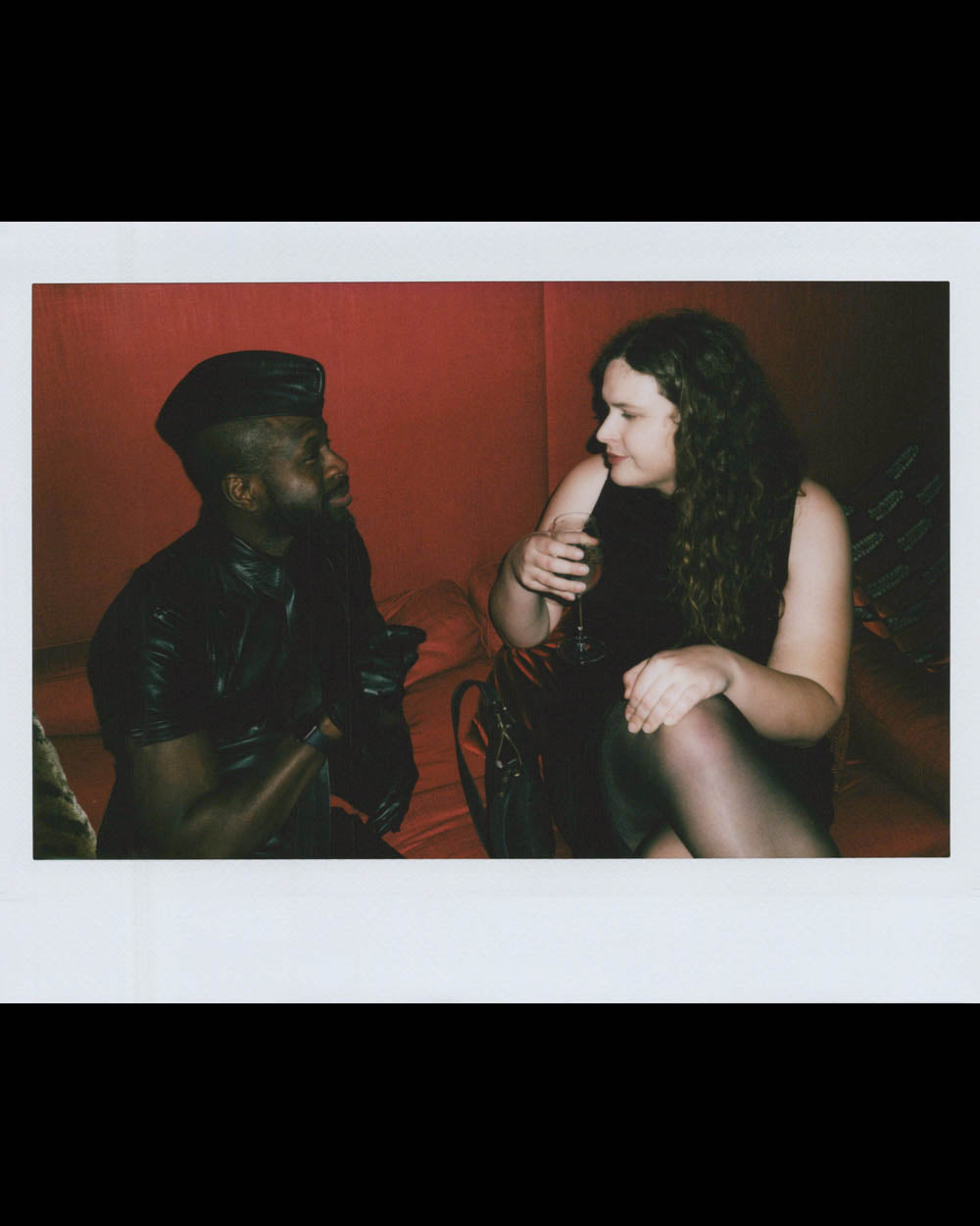 Man in black leather outfit and cap talking with woman holding a drink on red cushioned seating in low light.