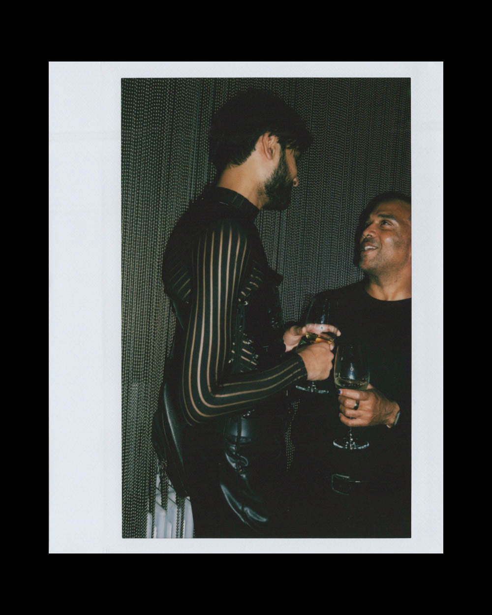 Two men sharing a drink and conversation, one wearing a striped sheer top and the other in black, against dark background.