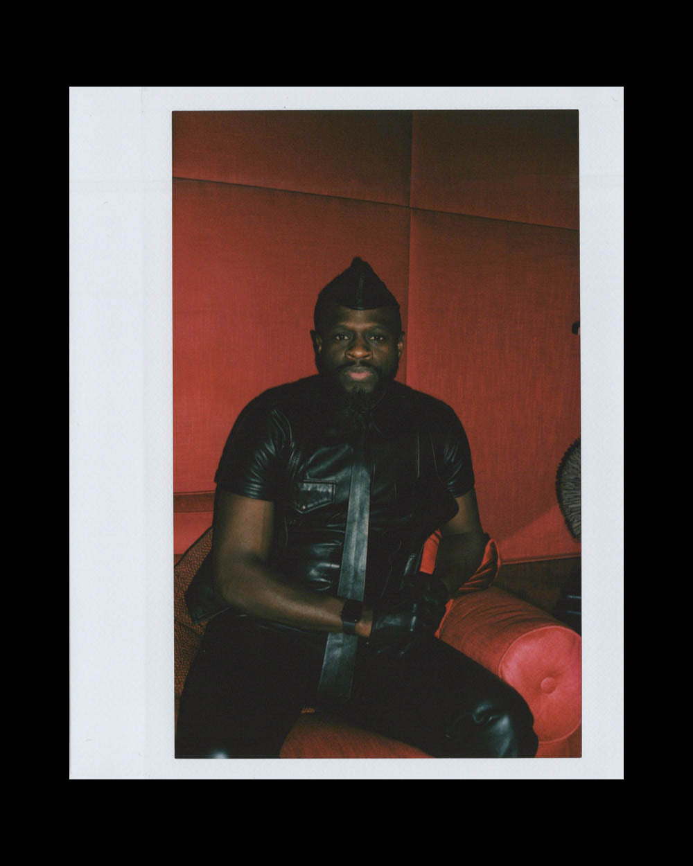 Man dressed in black leather outfit and matching gloves sitting on red upholstered seating against red wall backdrop
