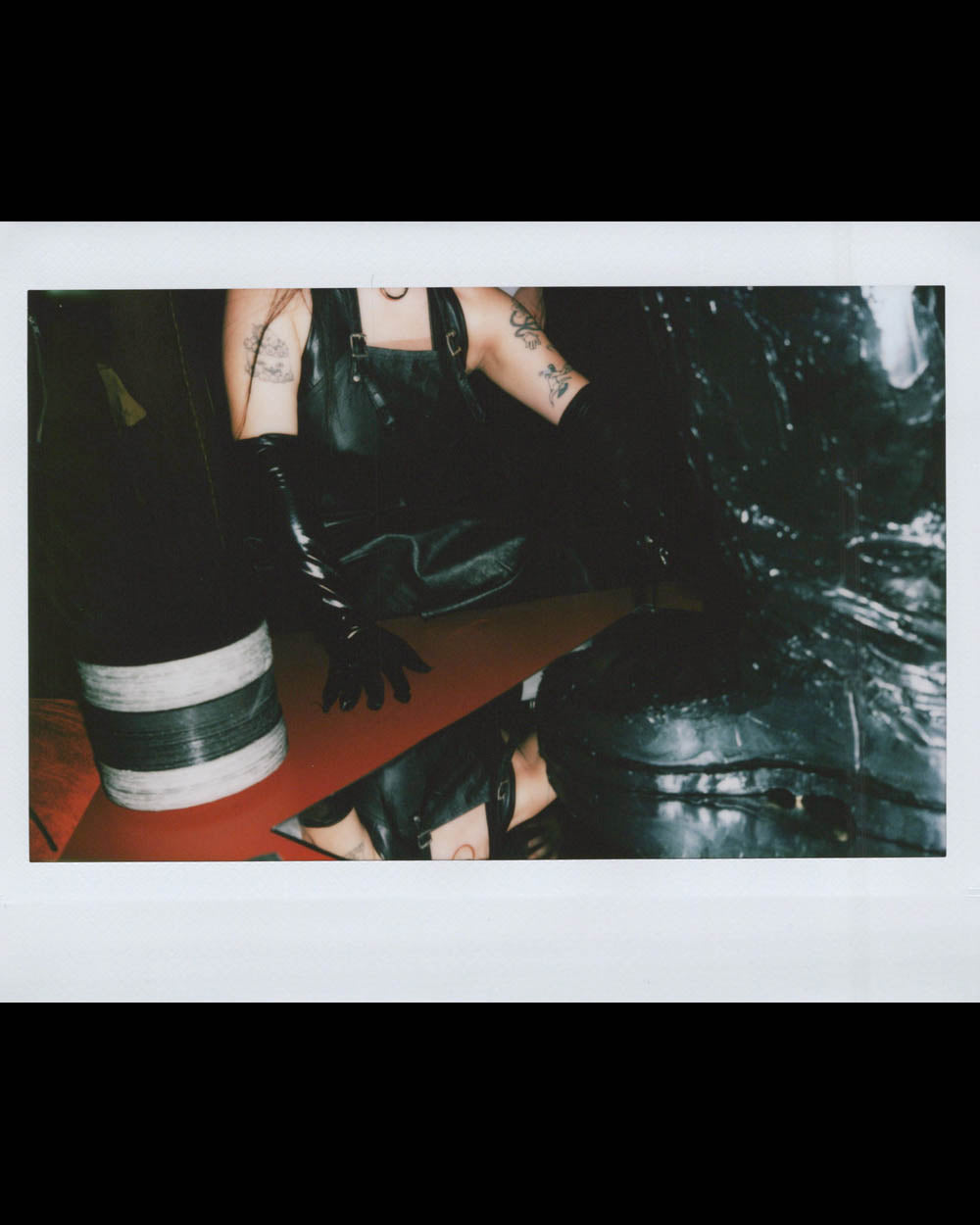 Person wearing long black gloves and leather apron sitting next to a reflective surface with tattoos visible on arms